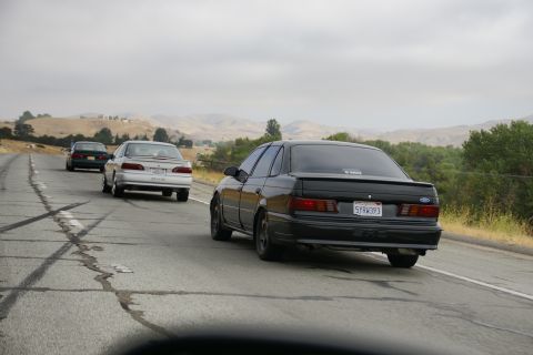 1995 Ford Taurus - Three sedans are driving on a highway with cracked pavement and a mountainous landscape.