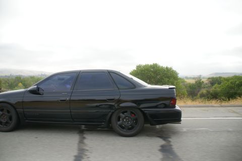1995 Ford Taurus - A black sedan is captured in a side profile shot while driving on a road.