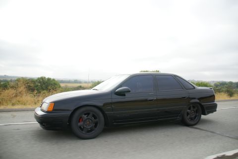 1995 Ford Taurus - A black sedan drives down a highway with a rural landscape in the background under an overcast sky.