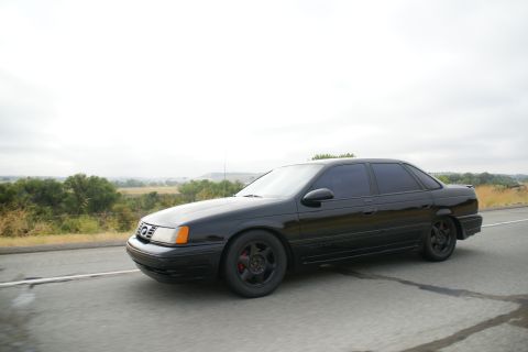 1995 Ford Taurus - A black 1995 Ford Taurus sedan is captured in a three-quarter front view while driving on a road with a scenic background.