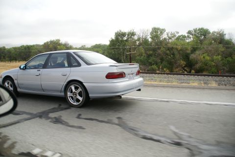 1995 Ford Taurus - A silver sedan is captured in motion on a highway, with railroad tracks visible alongside.