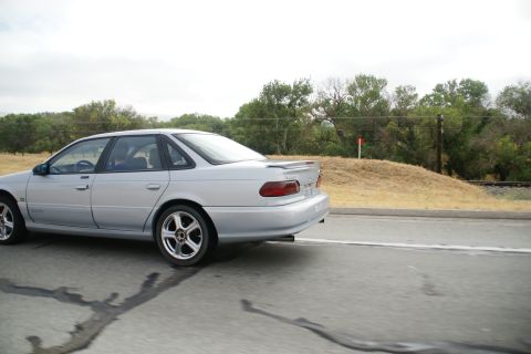 1995 Ford Taurus - A silver 1995 Ford Taurus sedan is captured from the side while driving on a road with trees and a railroad track in the background.