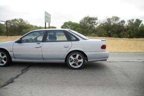 1995 Ford Taurus - A silver sedan is captured in motion on a highway, with trees and a sign visible in the background.