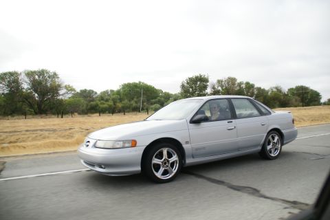 1995 Ford Taurus - Silver 1995 Ford Taurus sedan captured in motion on a highway under an overcast sky.