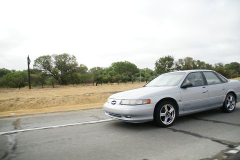 1995 Ford Taurus - A silver sedan is captured in motion on a highway, with a backdrop of trees and a cloudy sky.