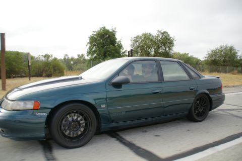 1995 Ford Taurus - A dark green 1995 Ford Taurus sedan with aftermarket black wheels is seen from the side.