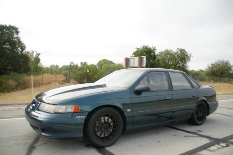 1995 Ford Taurus - A 1995 Ford Taurus sedan with a hood scoop and black wheels is seen driving on a road.