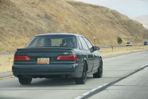 1995 Ford Taurus - A dark green sedan is seen from behind driving on a highway with dry hills in the background.