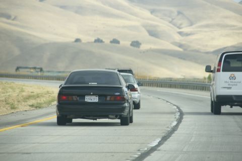 1995 Ford Taurus - A black 1995 Ford Taurus is seen driving on a highway with other vehicles and rolling hills in the background.