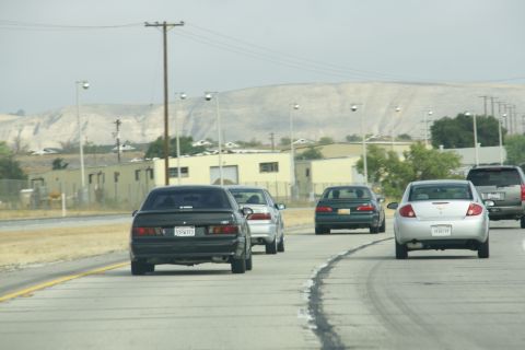 1995 Ford Taurus - Multiple vehicles travel down a highway under a hazy sky, with industrial buildings and barren hills visible.