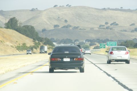 1995 Ford Taurus - A dark sedan drives on a highway with other vehicles and rolling hills in the distance.