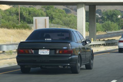 1995 Ford Taurus - Rear view of a black 1995 Ford Taurus sedan on a highway, with another car visible ahead.
