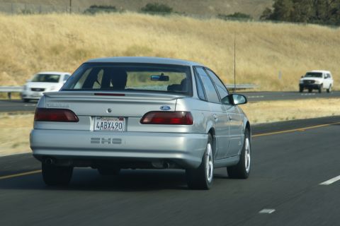1995 Ford Taurus - A silver 1995 Ford Taurus SHO is seen from behind while driving on a highway, with other vehicles in the background.