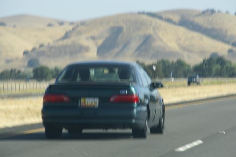 1995 Ford Taurus - A dark green sedan is seen from behind on a highway, with rolling hills in the distance.