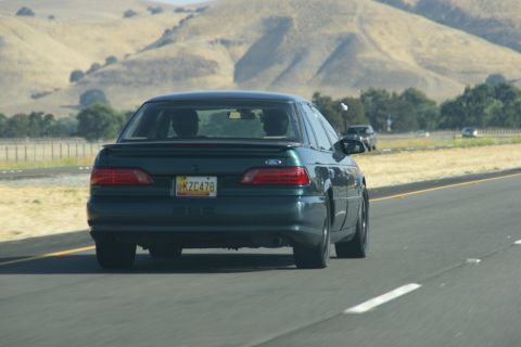 1995 Ford Taurus - A dark green 1995 Ford Taurus sedan is seen from the rear on a highway, with other vehicles in the distance.