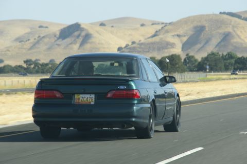 1995 Ford Taurus - Rear view of a 1995 Ford Taurus driving on a highway with dry hills in the background.