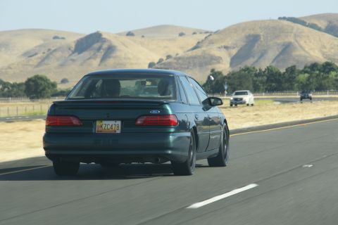 1995 Ford Taurus - A dark green 1995 Ford Taurus is seen from behind while driving on a highway with other vehicles and rolling hills in the distance.