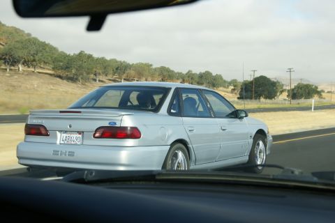1995 Ford Taurus - Silver 1995 Ford Taurus sedan seen from behind on a highway with trees and hills in the background.