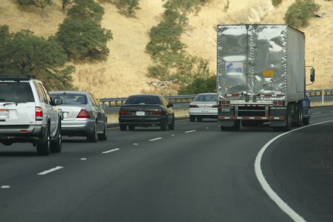 1995 Ford Taurus - A dark sedan is seen from behind on a highway, traveling alongside a large trailer truck and other vehicles.
