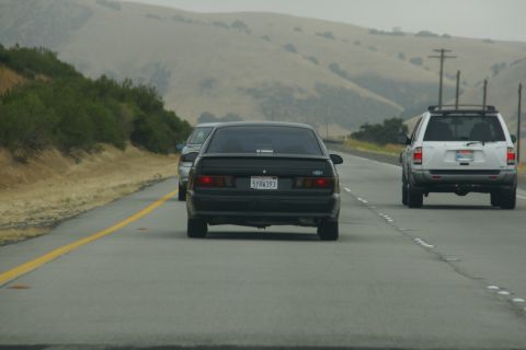 1995 Ford Taurus - A black sedan is seen from behind on a highway, with a white SUV in the adjacent lane and rolling hills in the distance.