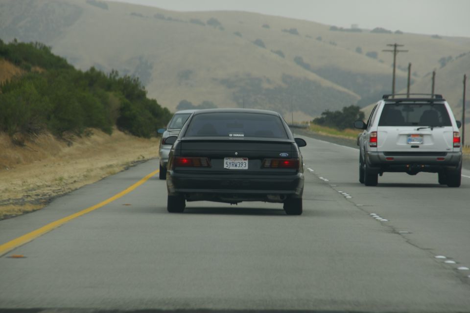 Black sedan driving on highway, white SUV in right lane, hills in background