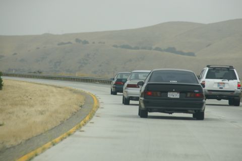 1995 Ford Taurus - A black 1995 Ford Taurus is seen from behind on a highway, with other vehicles and rolling hills in the background.