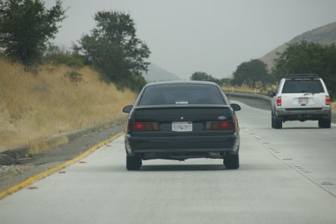 1995 Ford Taurus - A black sedan is seen from behind on a highway, with a white SUV in front of it.