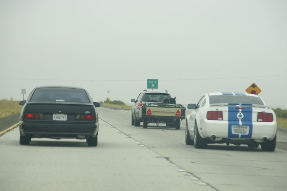 White Ford Mustang with blue stripes, black sedan, and pickup truck towing a trailer on highway