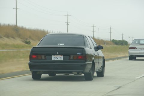 1995 Ford Taurus - A black sedan is seen from behind driving on a highway, with another vehicle in front of it.