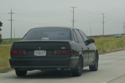 1995 Ford Taurus - Rear view of a black 1995 Ford Taurus sedan driving on a highway under an overcast sky.