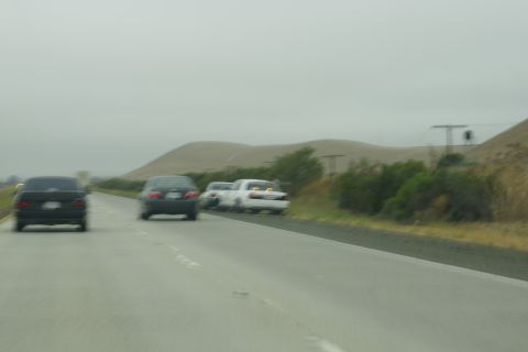 1995 Ford Taurus - Three cars are seen from behind on a highway with rolling hills in the background under a cloudy sky.