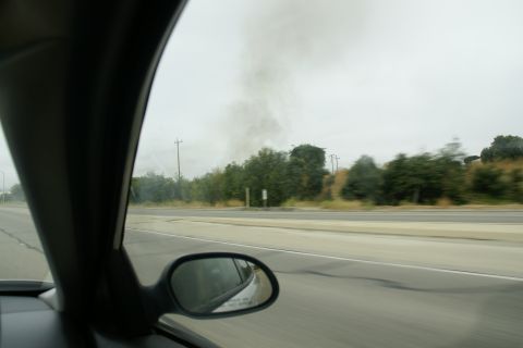 1995 Ford Taurus - A plume of dark smoke rises into the overcast sky, visible from the passenger side of a moving vehicle.