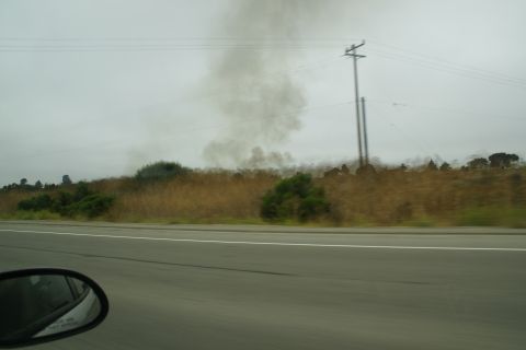 1995 Ford Taurus - Smoke billows into the sky from a field beside a highway on an overcast day.
