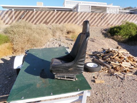 1982 Ford Bronco - A black bucket car seat sits on a green board outdoors next to a pile of wood and a spare tire.