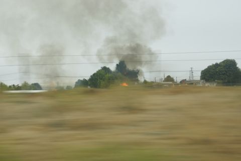 1995 Ford Taurus - A plume of dark smoke rises into the sky, obscuring a distant fire and industrial structures.