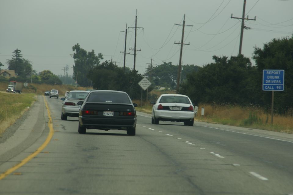 black sedan and white sedan driving on a two-lane road, with a sign that says 'REPORT DRUNK DRIVERS CALL 911'