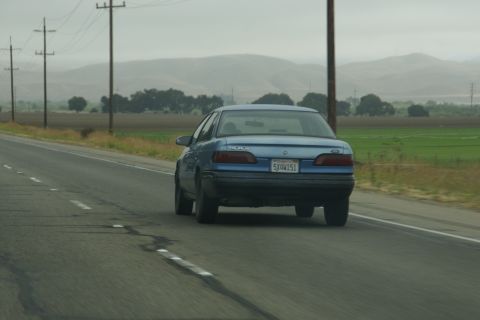 1995 Ford Taurus - A blue sedan travels down a highway with fields and hills in the background under an overcast sky.