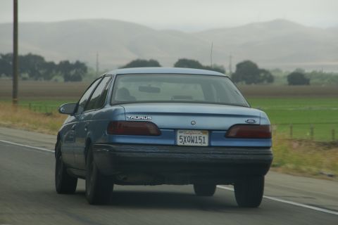 1995 Ford Taurus - A blue 1995 Ford Taurus sedan is seen from behind on a highway with rolling hills in the background.