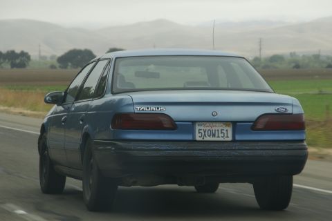 1995 Ford Taurus - Rear view of a blue 1995 Ford Taurus driving on a highway, showing its distinctive taillights and California license plate.