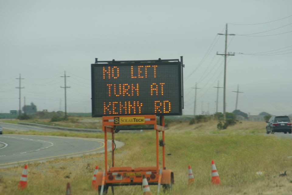 Digital road sign displaying "NO LEFT TURN AT KENNY RD" with a dark SUV in the background