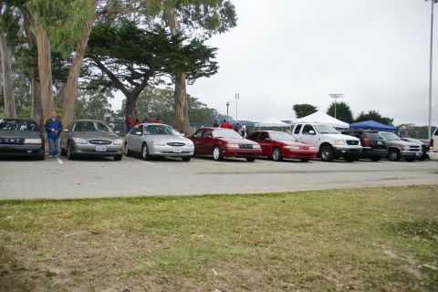 1995 Ford Taurus - A variety of vehicles are parked in a line under an overcast sky, with trees and tents visible.