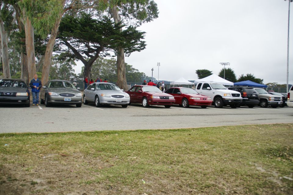 Several cars and trucks parked in a row, with trees and tents in the background