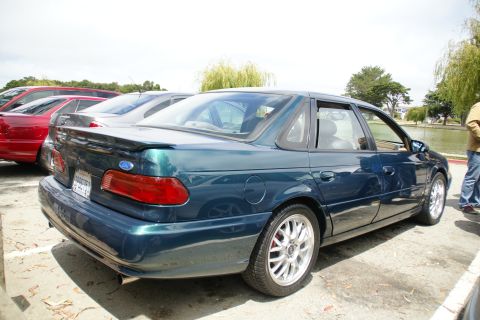 1995 Ford Taurus - A dark green sedan with a spoiler is parked in a lot near a body of water.