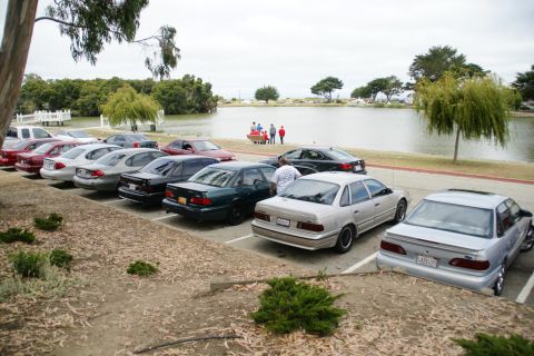 1995 Ford Taurus - Several sedans are parked in a lot next to a lake on an overcast day.