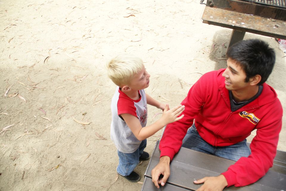 Boy in red and gray shirt and jeans touches man in red hoodie and jeans at picnic table