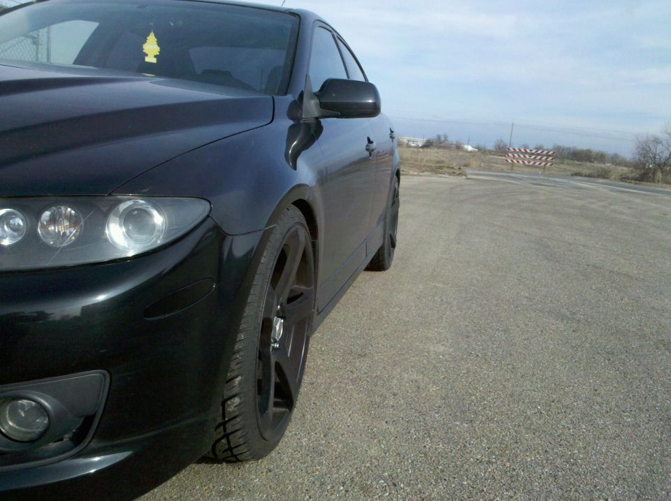 Black sedan parked on asphalt, front wheel and fender visible. Sky is overcast