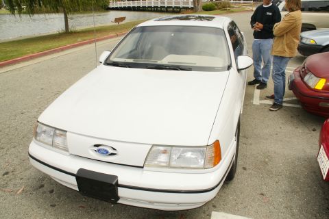 1995 Ford Taurus - A white 1995 Ford Taurus sedan is parked next to other vehicles, with two people standing nearby.