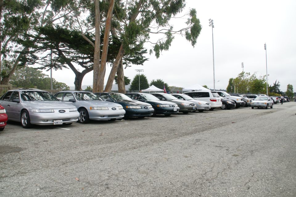 Line of sedans and SUVs parked in a lot, with trees and tents in the background. The first car is a silver 1995 Ford Taurus
