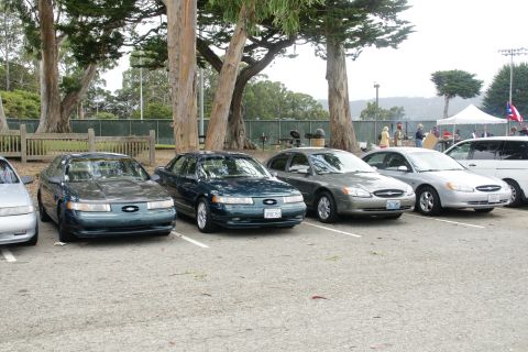 1995 Ford Taurus - Several sedans are parked in a lot with trees and people in the background.