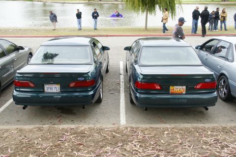 1995 Ford Taurus - Two dark green Ford Tauruses are parked in a lot near a lake, with people in the background.